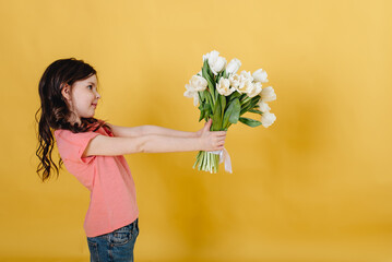 Photo of a cute girl on a yellow background holding a bouquet of white tulips as a gift that she will give to her mother 
