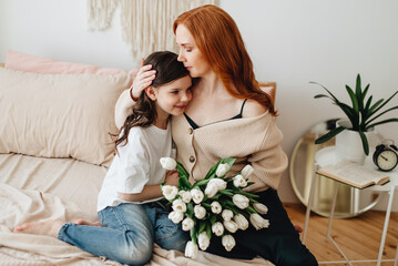 Happy beautiful affectionate mother, with a bouquet of spring flowers, hugs her daughter at home on the bed, enjoys the moment of surprise, celebrates Mother's Day.