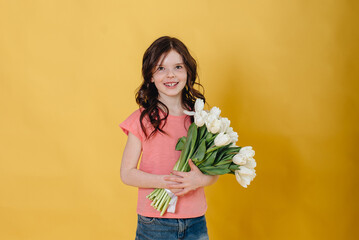 Photo of a cute girl on a yellow background holding a bouquet of white tulips as a gift that she will give to her mother