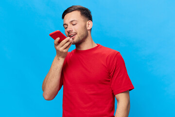 Smiling happy tanned handsome man in red t-shirt record voice message to friend using phone posing isolated on blue studio background. Copy space Banner Mockup. Online People lifestyle concept