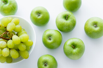 top view of green and ripe apples near grapes in bowl on white.