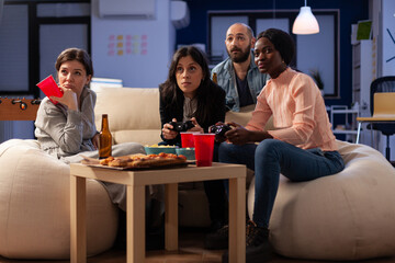 Diverse group of coworkers having fun with video games on console, enjoying drinks and snacks at office celebration after work hours. People using controller to play online game at gathering.