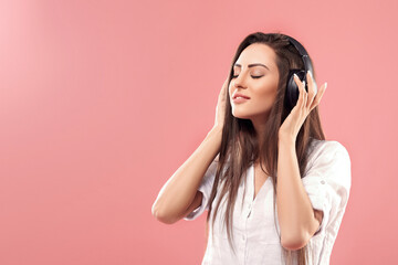 Beautiful young woman in wireless headphones listening to music and dancing on pink background. Girl uses wireless earphones.