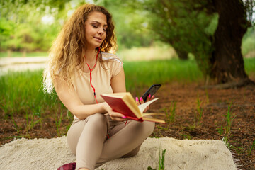 Obraz premium Image of positive caucasian woman sitting outdoors in park on a grass and using mobile phone while listening music. she holding book in other hand.