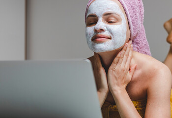 Gorgeous young adult woman in cosmetics facial mask touching neck with her hands and looking at laptop.
