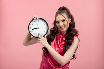 Smiling young brunette female girl in pink blouse posing isolated on pastel pink background in studio. People sincere emotion lifestyle concept. Pointing with index finger on clock.