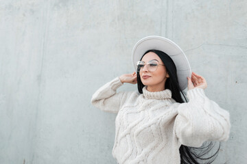 Happy beautiful Caucasian woman with a lovely face and glasses wearing a fashionable knitted sweater adjusts her hat against a gray grunge concrete background