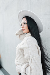 Stylish beautiful young woman with long brunette hair and trendy glasses in a fashionable white hat with a knitted vintage sweater walks on the street near the concrete wall