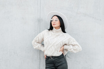 Happy beautiful young girl with fancy vintage glasses and fashionable clothes with a sweater, hat, and leather pants stands near a concrete gray wall on the street