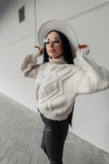 Happy Caucasian woman with smile and vintage glasses in trendy beige knit sweater and hat walks in the city