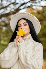 Happy beautiful young girl model with a smile in a fashionable vintage clothing with a hat and sweater holding a yellow autumn leaf near her face and walking in the park