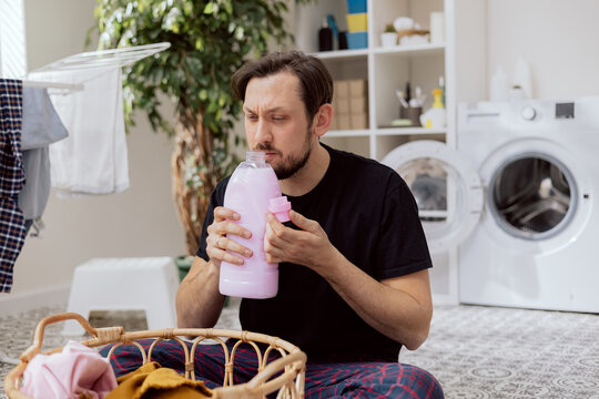 Laundry Room, Bathroom. Man Dressed In Household Clothes Sits On Floor By Wicker Basket With Things To Wash. Man Holds Laundry Detergent In Hands And Sniffs Its Scent. A Wry Face, He Doesn't Like It.