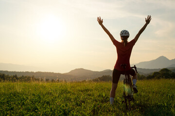 Woman on a road racing bike stopping at a green glade, celebrating with arms raised while looking...
