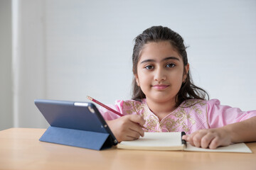 portrait cute Indian girl writing on notebook and online learning class from tablet, education...