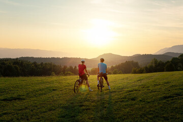 Naklejka na ściany i meble Woman and man, road racing bikers, sitting on bicycles, enjoying a beautiful mountain landscape at sunset