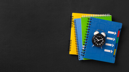 Obraz premium A stack of colored school notebooks and an alarm clock on a school chalkboard background. Back to school concept, banner