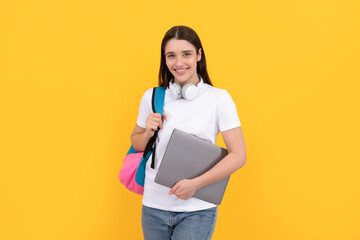 young girl with backpack hold laptop in headphones on yellow background, education