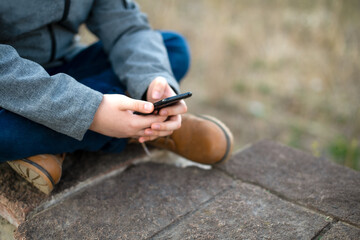 A schoolboy boy uses a mobile phone in his hands in nature in the city in the spring. Free time.
