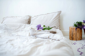 Fototapeta na wymiar Bed with beautiful white crumpled linenlinens in a white bedroom and Lilac flowers. Working office work remotely from home on bed. Distance learning online education and work.