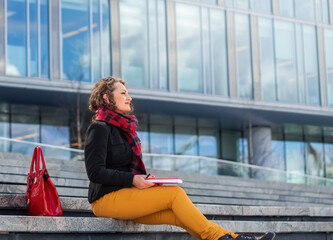 Attractive business woman sits on the steps during a break with a notebook and a pen in her hands against the backdrop of a business center and looks into the distance smiling