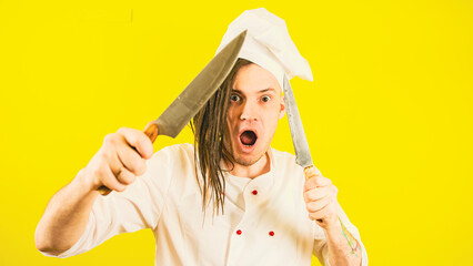 Young insane man dressed as chef with knives on yellow background. Male cook in white hat and shirt posing with kitchen knives.