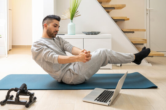 Young Guy With Average Body Type Trying To Improve His Muscles, Lose Belly Fat And Weight On Specific Body Parts While Watching Online Workout Courses And Doing Russian Twist On Mat At Home.