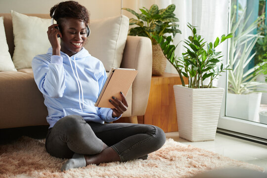 Happy young woman in comfy clothes sitting on floor at home and watching short funny videos on tablet computer