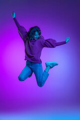 Full-length portrait of young excited girl, student jumping isolated over purple background in neon. Concept of emotions, facial expression, art, beauty