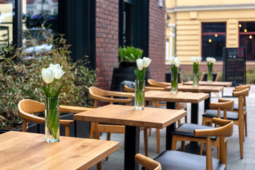 Fototapeta na wymiar Empty tables with flowers in outdoor cafe or restaurant. Tables and chairs at sidewalk cafe. Touristic setting, tulips on cafe table, sidewalk cafe furniture.