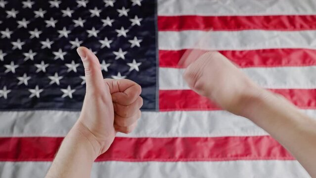 thumbs up and thumbs down gesture signals made with two caucasian hands in front of blurry US flag background