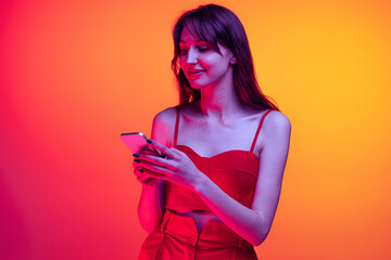 Portrait of young emotive girl, student using phone isolated over orange studio background in neon...