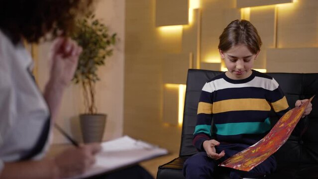 Preteen boy holding his drawing and explaining artwork to child counselor during psychology session. Female therapist making notes analyzing patient's artwork during consultation