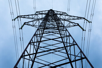 View from the bottom upwards to steel tower of electric main or electricity transmission line with the wires silhouette on background of cloudy blue sky
