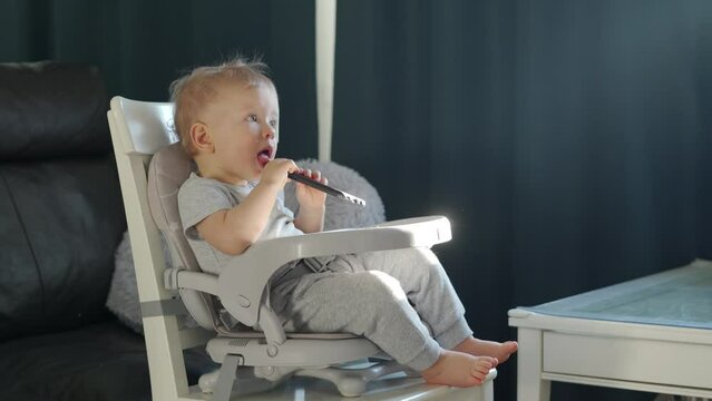 Cute kid sitting in booster seat fixed on top of dining chair, baby playing with mobile phone. High quality 4k footage