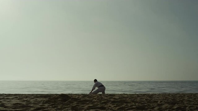 Sporty man doing warm-up squatting on sand. Karate fighter stretching on beach.