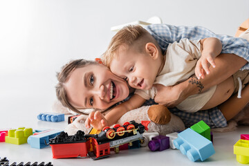 cheerful woman looking at camera while hugging toddler son near toys on white.