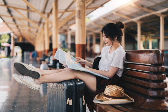 Happy young asian woman traveler or backpacker using map choose where to travel with luggage at train station, summer vacation travel concept