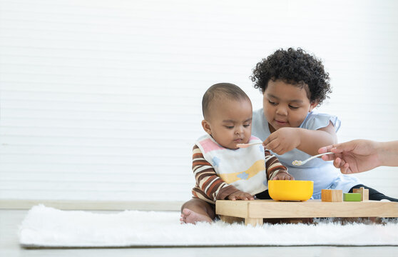 Little cute African older sister helping mother to feeding her newborn sister food while baby playing toys sitting on floor at home. Relationship of Siblings concept. White background. Copy space