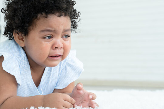Side view of little sad African American chubby kid girl is crying with tears rolled down her cheeks while lying on fluffy carpet on floor at home. Child emotion care concept. White background