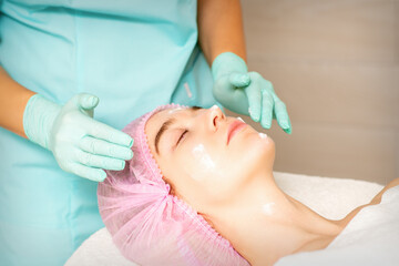 Fototapeta premium Cosmetologist with gloved hands applies a moisturizing mask with peeling cream on the female face. Facial cosmetology treatment. Procedures for facial care