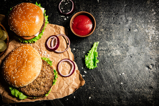 Burger on a cutting board with onion rings, tomato sauce and lettuce. On a black background. 
