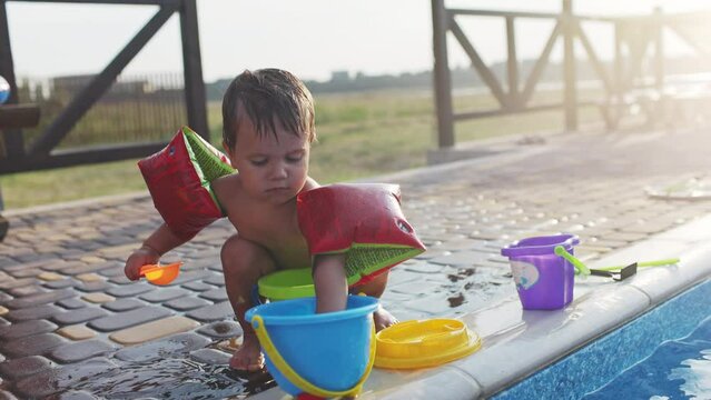 Boy with armbands playing with toys near the pool with clear water on the background of a summer sunset