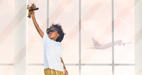 African american boy wearing googles flying toy airplane and flying airplane seen through window