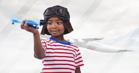 African american boy with toy plane wearing aviator hat and goggles and airplane flying against sky - Powered by Adobe