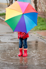 Obraz premium A cute little girl in a red cape, red boots and a white hat jump in puddles and has a fun.The girl has a rainbow umbrella in her hands. Happy childhood. Early spring. Emotions.