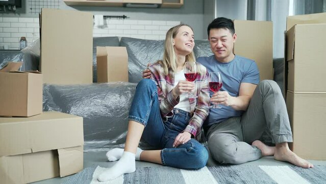 Young married couple celebrating moving to a new apartment. Buying a home. Happy family husband and wife drinking wine between cardboard boxes. Happiness own housing. Toasting in their renovated house