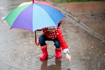 A cute little girl in a red cape, red boots and a white hat jump in puddles and has a fun.The girl...