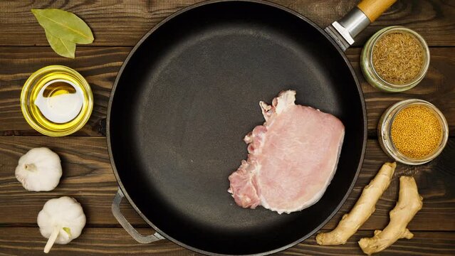 The cook puts raw meat steaks in a frying pan. Sliced pork meat in a cast iron skillet. Fry ingredients. Olive oil, bay leaf, garlic, mustard seeds, zira, ginger root, dry spices. Close-up. Top view