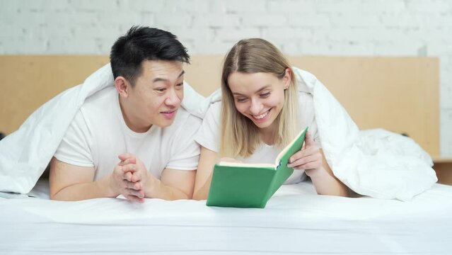 Happy young multiracial family reading a book together lying in bed. Mixed race couple spends leisure time together on weekends in the morning. Cheerful Asian husband and wife in bedroom at home