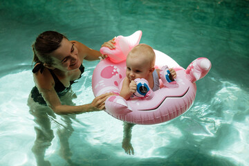 Young attentive mother is helping her little daughter swim in turquoise water during swimming class...